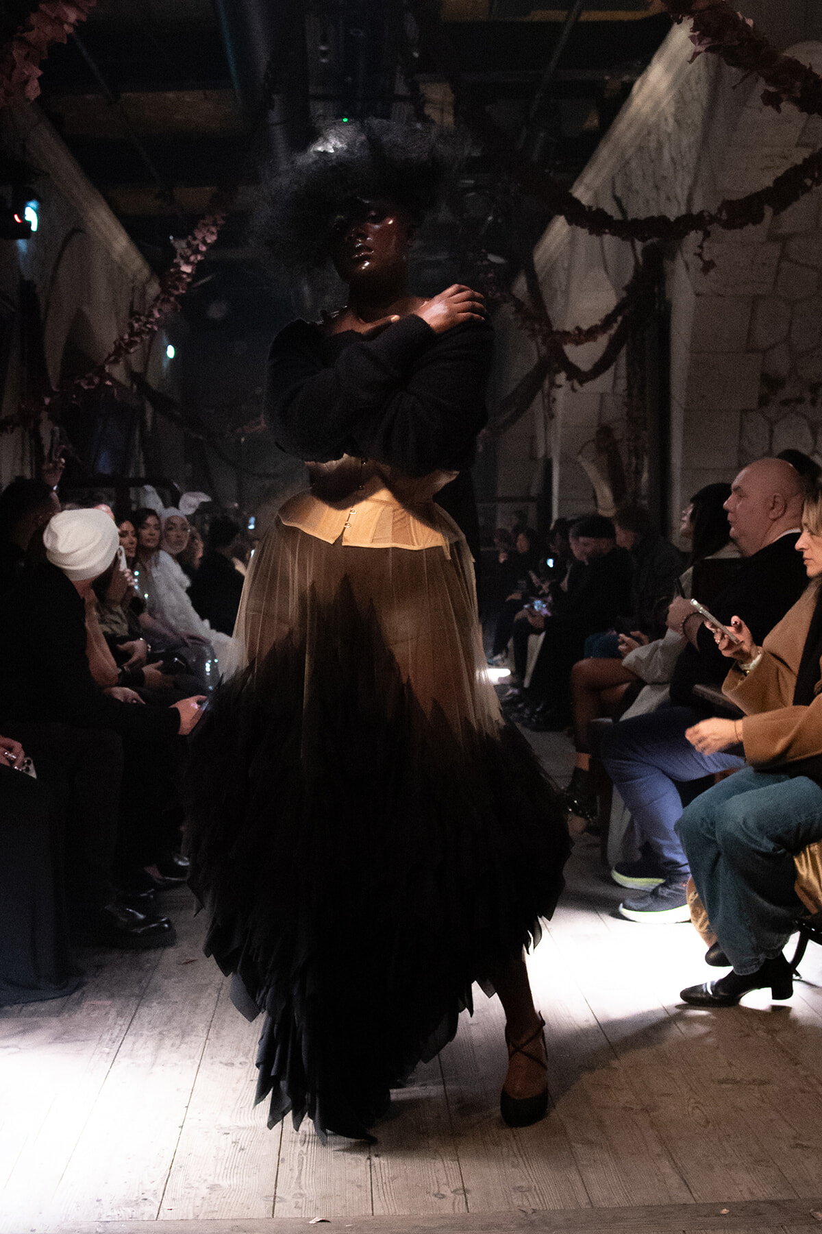 Model walks a dimly lit runway in a black top and a voluminous beige-to-black layered tulle skirt; audience seated along the sides watching.