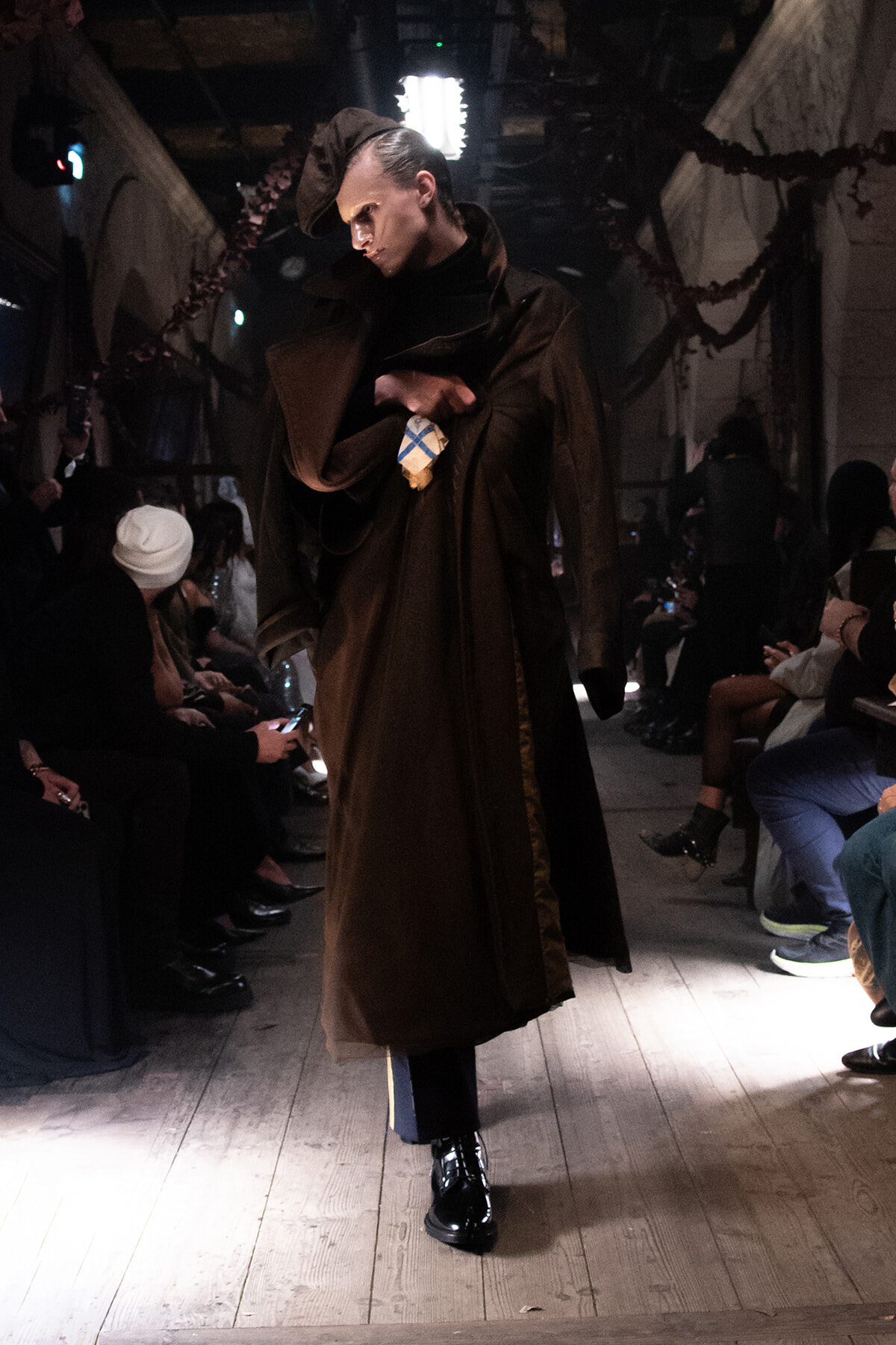 Model walking the runway in a long brown cloak and matching hat, spotlight on a wooden-floor runway with seated audience on both sides.