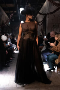 Model walking the runway in a dark brown sheer gown with a corset-like waist, surrounded by seated audience taking photos in a dim venue.