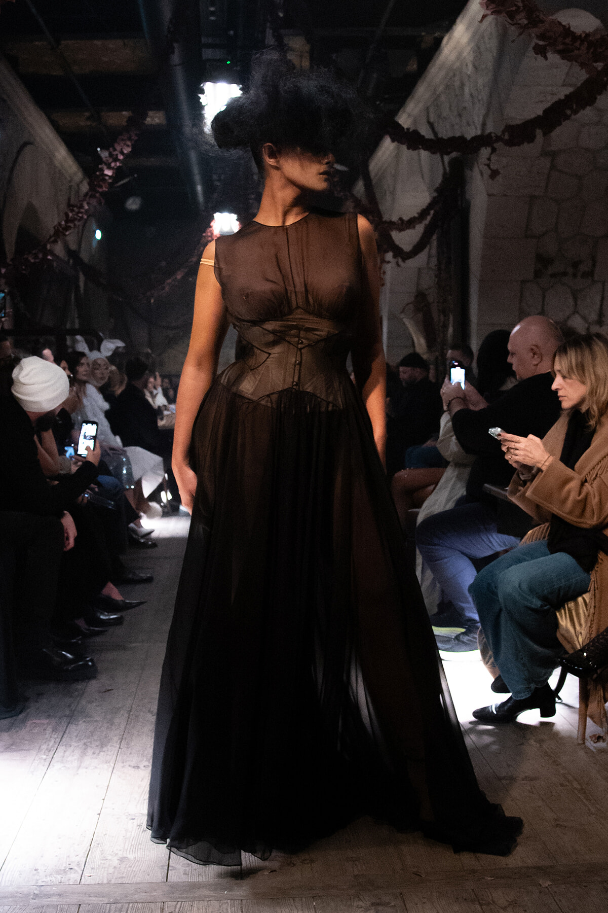 Model walking the runway in a dark brown sheer gown with a corset-like waist, surrounded by seated audience taking photos in a dim venue.