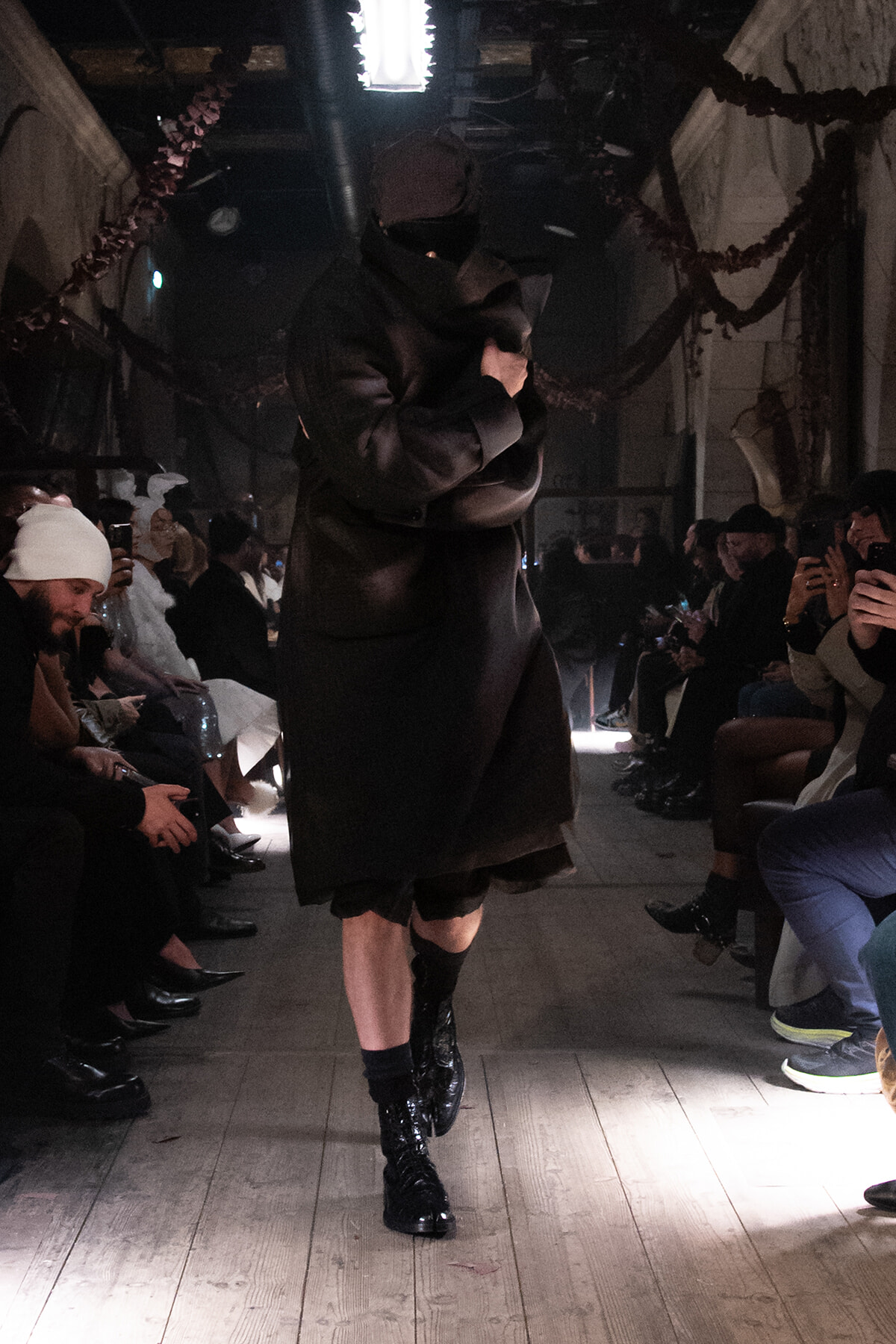 Model walks the runway in a dark oversized black coat and glossy boots as seated spectators photograph the scene under dim lighting.