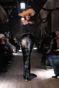 Model walking a fashion runway in a long, high-gloss black skirt and cropped dark top, audience watching on either side.