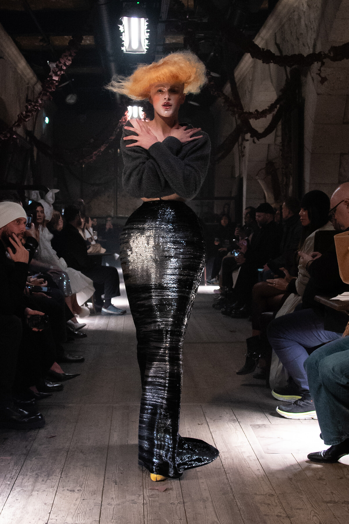 Model walking a fashion runway in a long, high-gloss black skirt and cropped dark top, audience watching on either side.