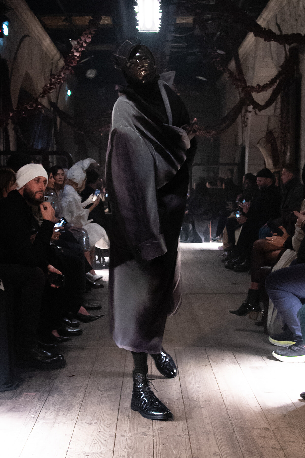 Model walks the runway in a metallic, flowing gray-black outfit with a glossy dark mask, as spectators sit along both sides.