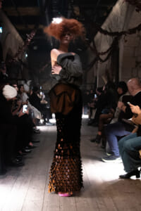 Model walking a runway in an off-shoulder gray sweater and a long skirt with circular cutouts; audience on both sides.