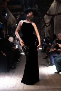 Model in a sleek black gown with circular side cut-outs walks the runway as spectators film with their phones.