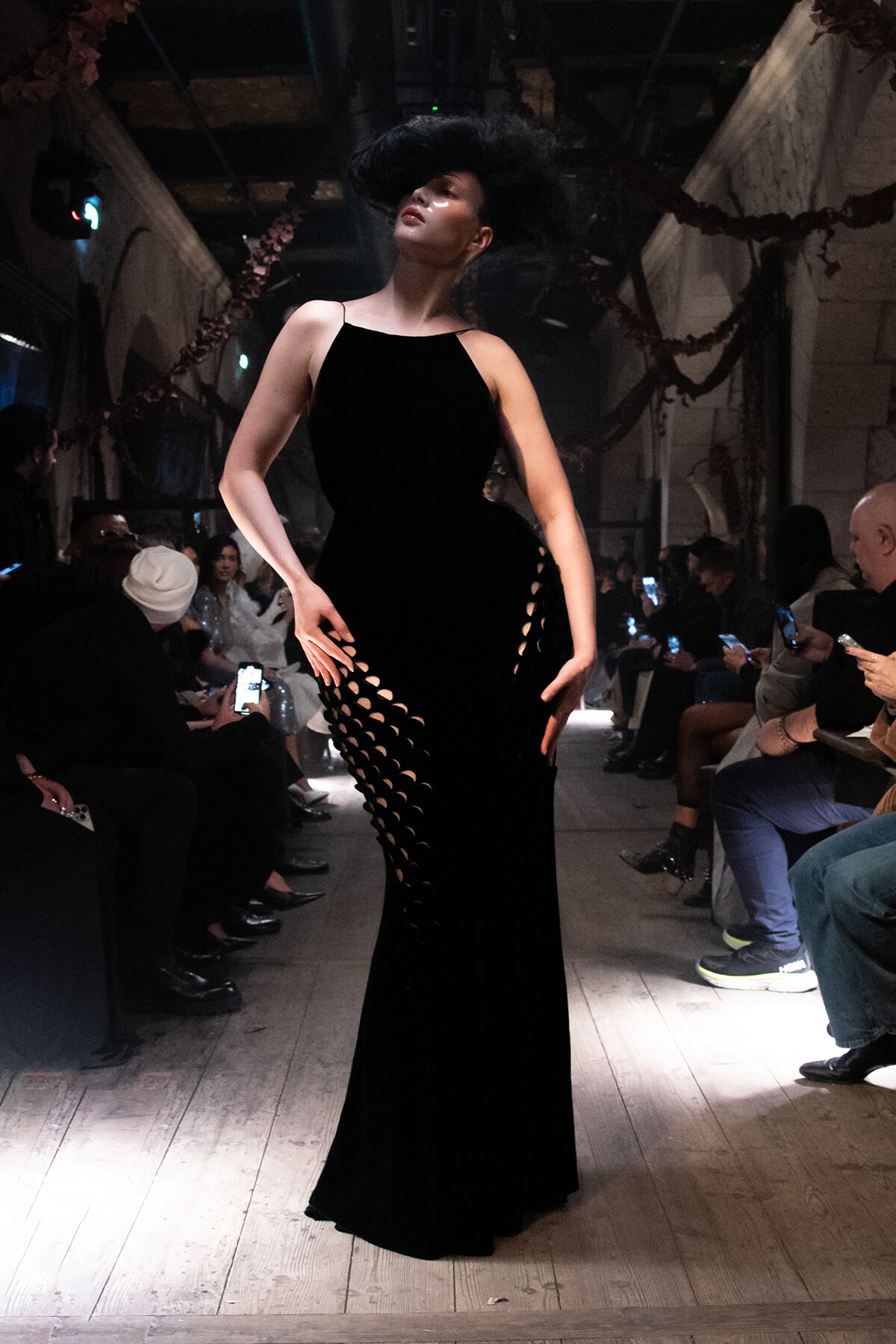 Model in a sleek black gown with circular side cut-outs walks the runway as spectators film with their phones.
