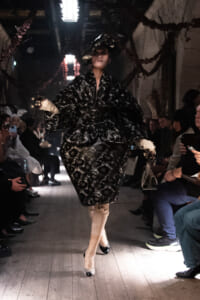 Model walks the runway in a black lace voluminous coat with beige boots as seated audience watches on both sides.