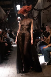 Model on a runway wearing a dark sheer lace gown with a high neckline and flowing skirt, red hair obscuring her face, audience on both sides in dim lighting.