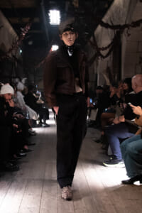Model walking a fashion runway in a dark brown denim jacket, black scarf, cap, and leopard-print shoes, audience seated along wooden floor runway in dim venue.