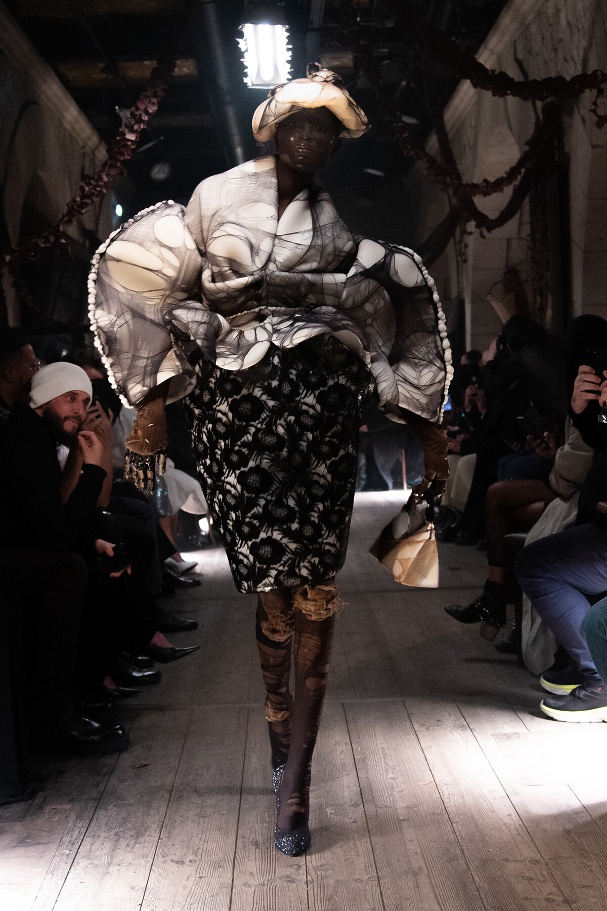 Model walks a runway in a dramatic black-and-white patterned dress with oversized sculptural sleeves and a white hat; audience watches from both sides.
