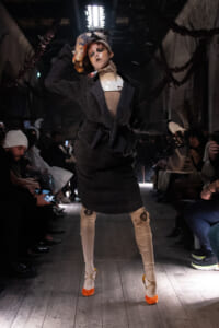 Model on a dimly lit runway wearing a dark oversized coat, distressed beige pants, and bright orange heels, posing to the audience.