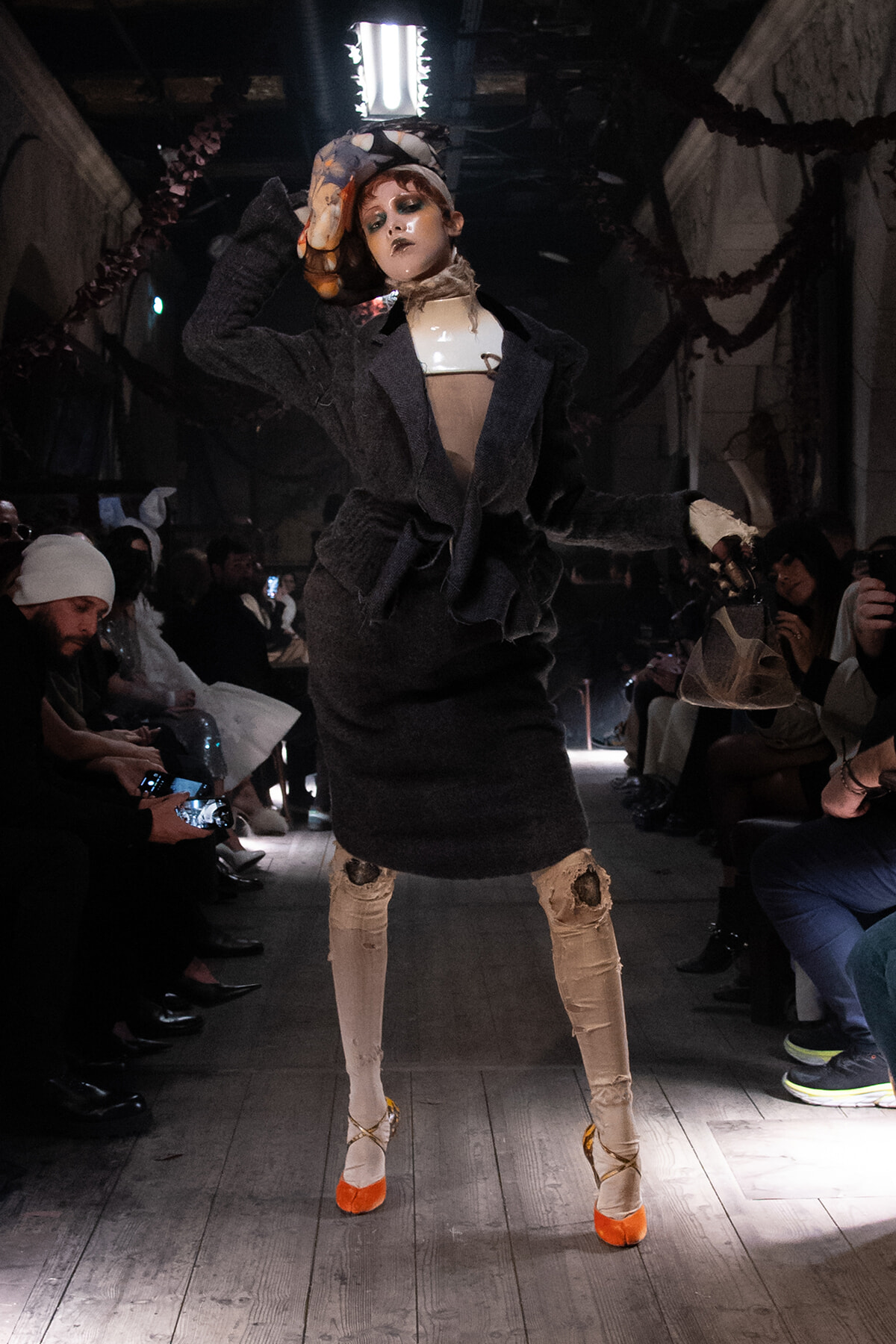 Model on a dimly lit runway wearing a dark oversized coat, distressed beige pants, and bright orange heels, posing to the audience.