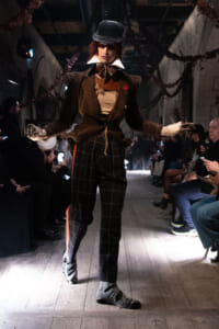 Model walks a runway in a vintage-inspired brown tweed suit with white collars, plaid pants, hat, and gloves as spectators watch from the sides.