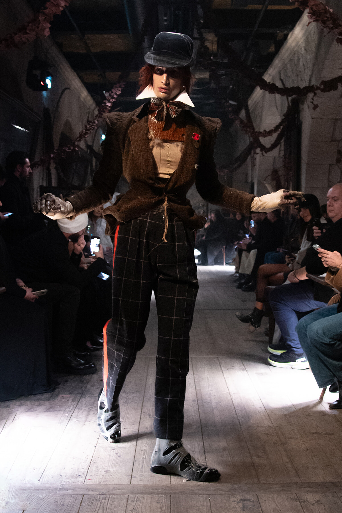 Model walks a runway in a vintage-inspired brown tweed suit with white collars, plaid pants, hat, and gloves as spectators watch from the sides.