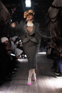 Model walking a fashion runway in a gray textured suit with a large beige hat and bright pink socks, arms extended as she poses.
