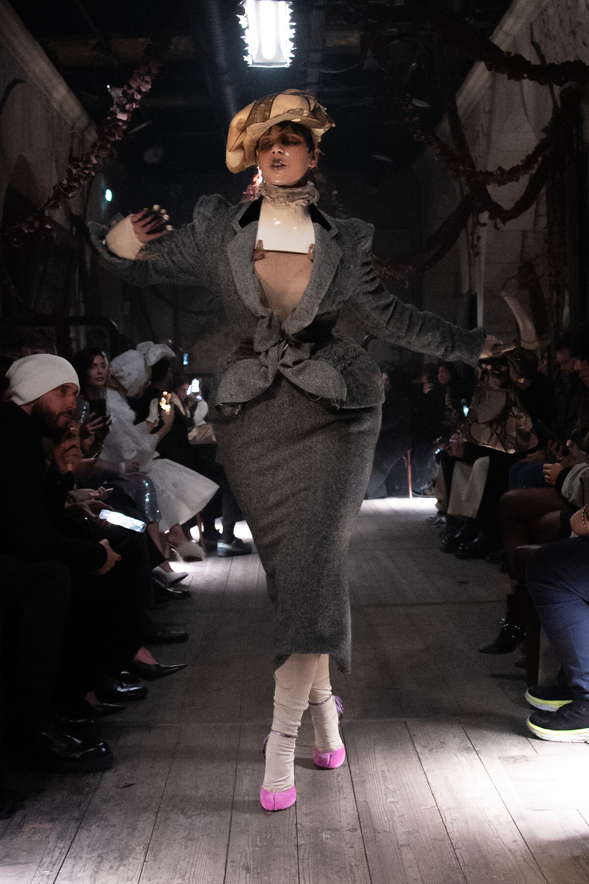 Model walking a fashion runway in a gray textured suit with a large beige hat and bright pink socks, arms extended as she poses.
