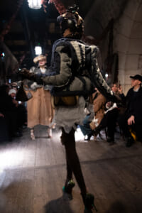 Model on a runway wearing a patchwork, distressed costume with large shoulder accents, viewed from behind as audience watches.