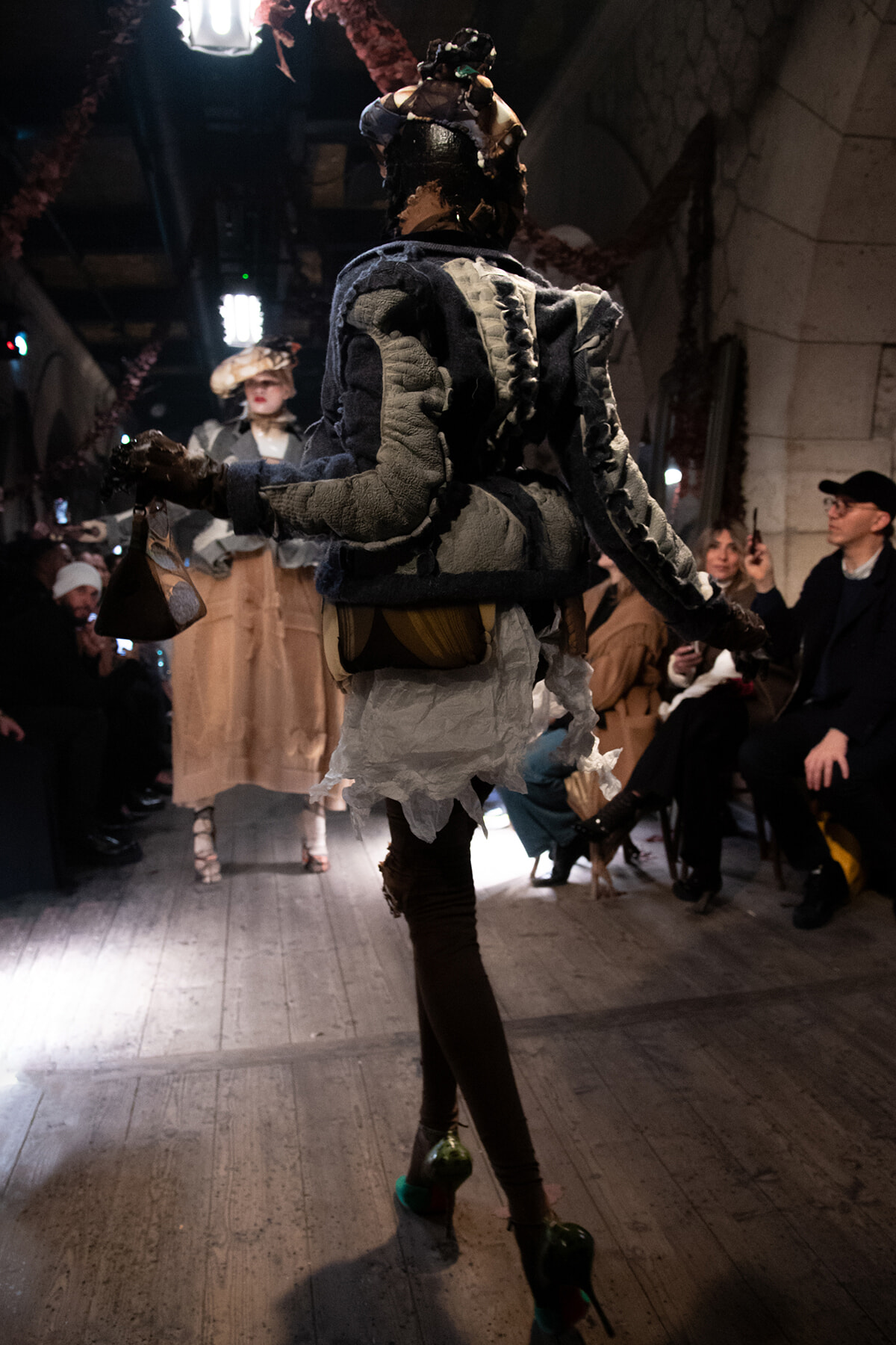 Model on a runway wearing a patchwork, distressed costume with large shoulder accents, viewed from behind as audience watches.