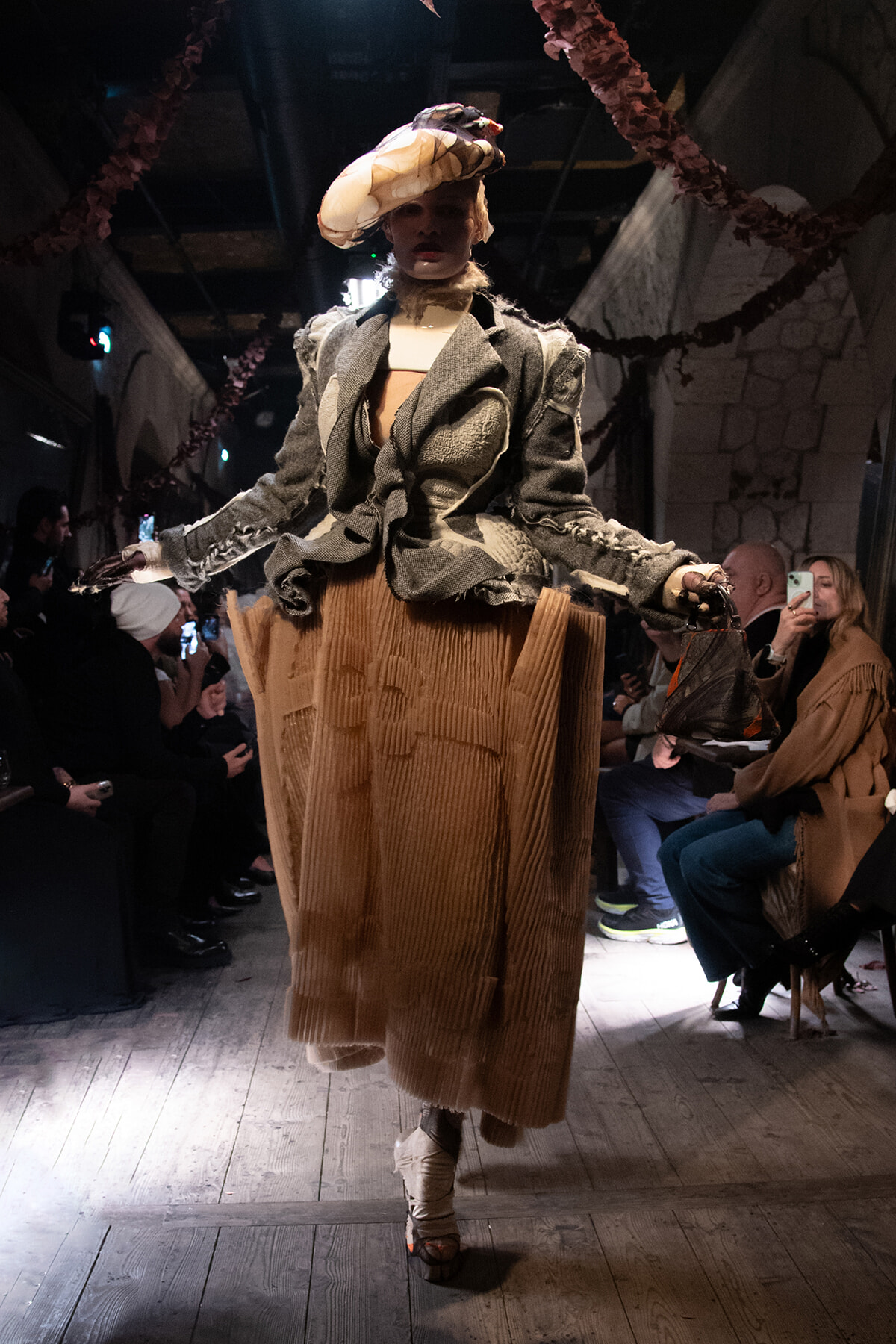 Model walking a runway in a textured gray blazer and tan pleated skirt with an oversized hat, audience watching from both sides.