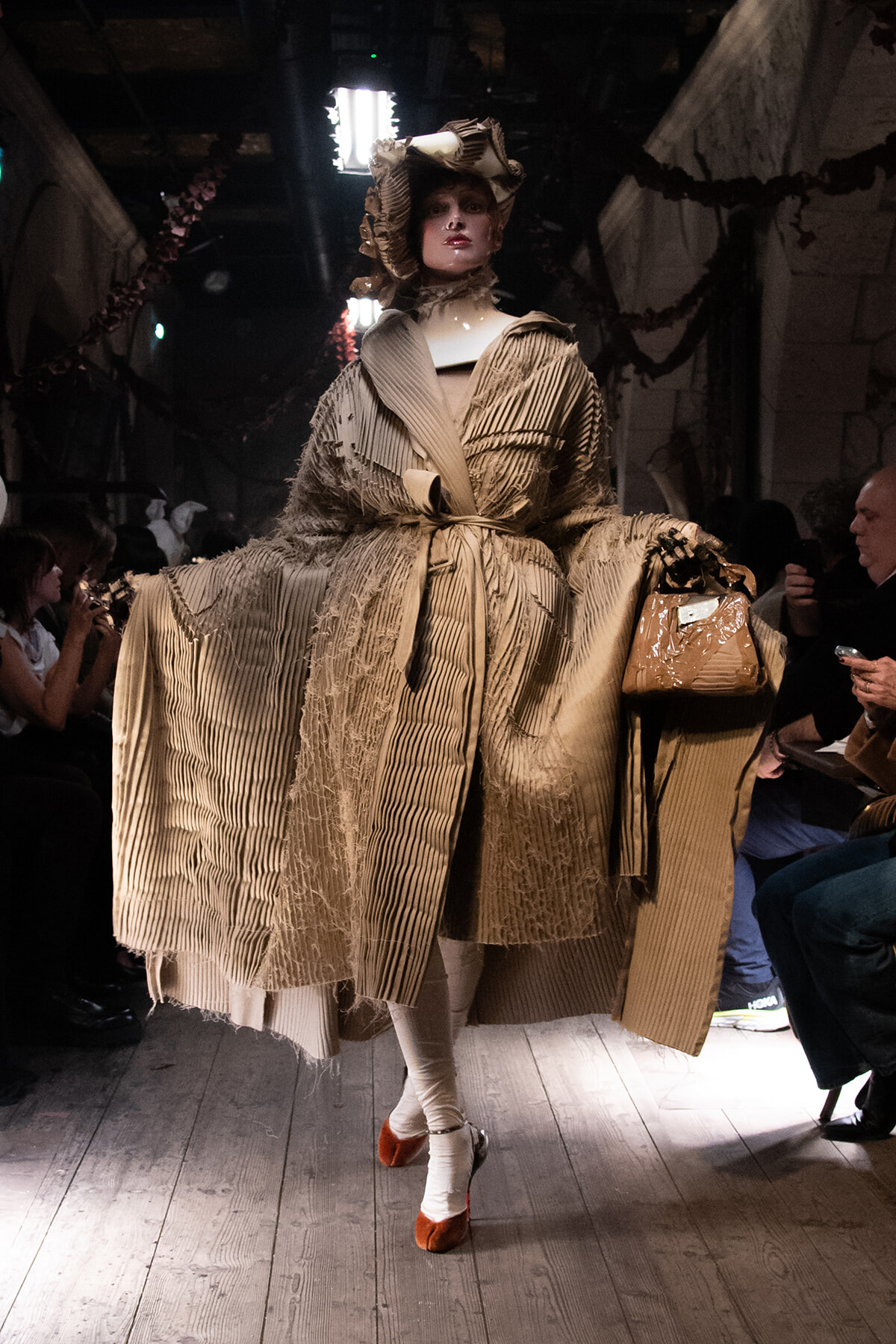 Model walks the runway in a large beige pleated gown with a sculptural hat and tan handbag.