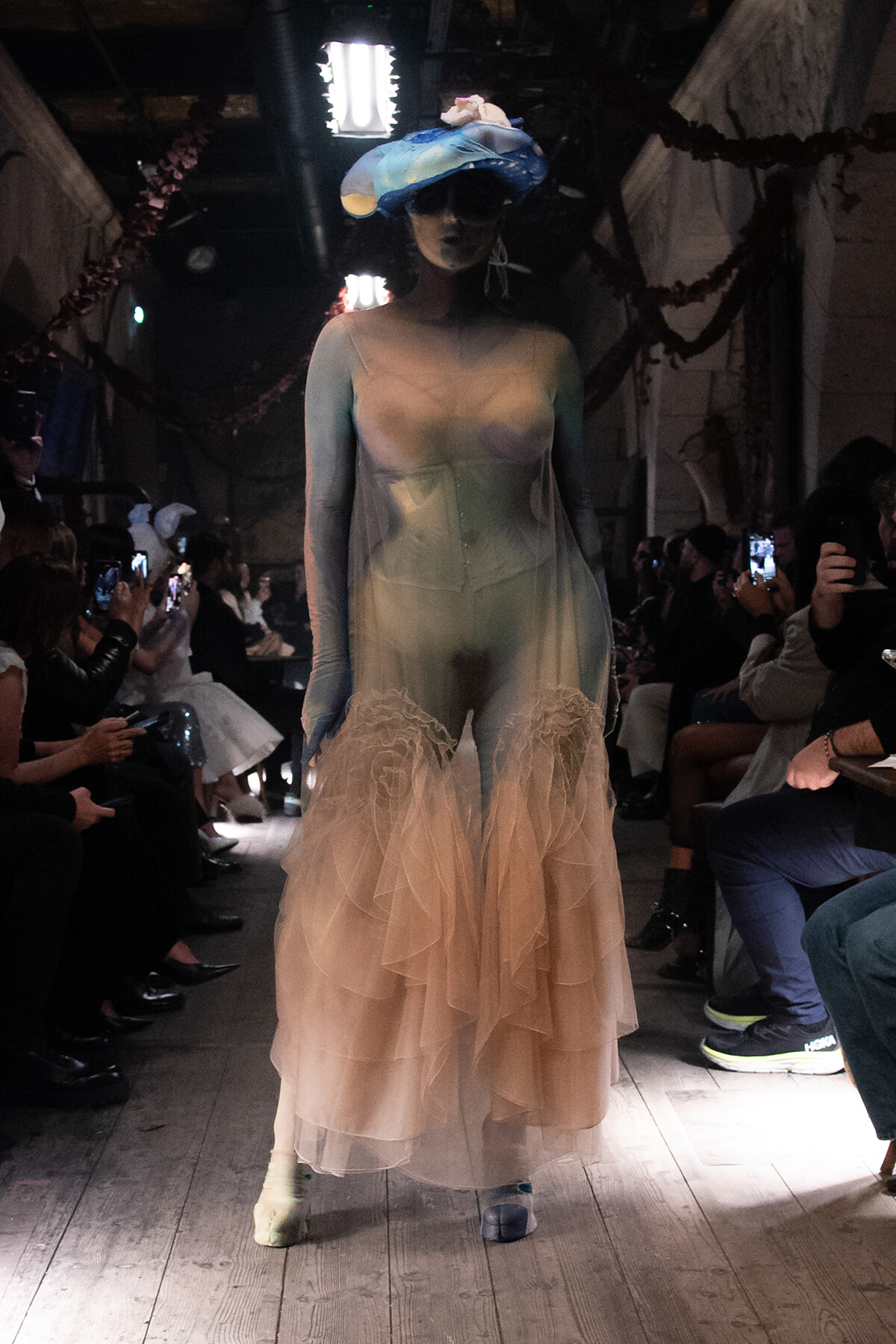 Model walks the runway in a sheer gradient gown with layered tulle; oversized blue headpiece with pink accents, audience on both sides taking photos.