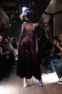Model walks the runway in a gradient purple dress with a large colorful flower headpiece, audience watching from both sides.