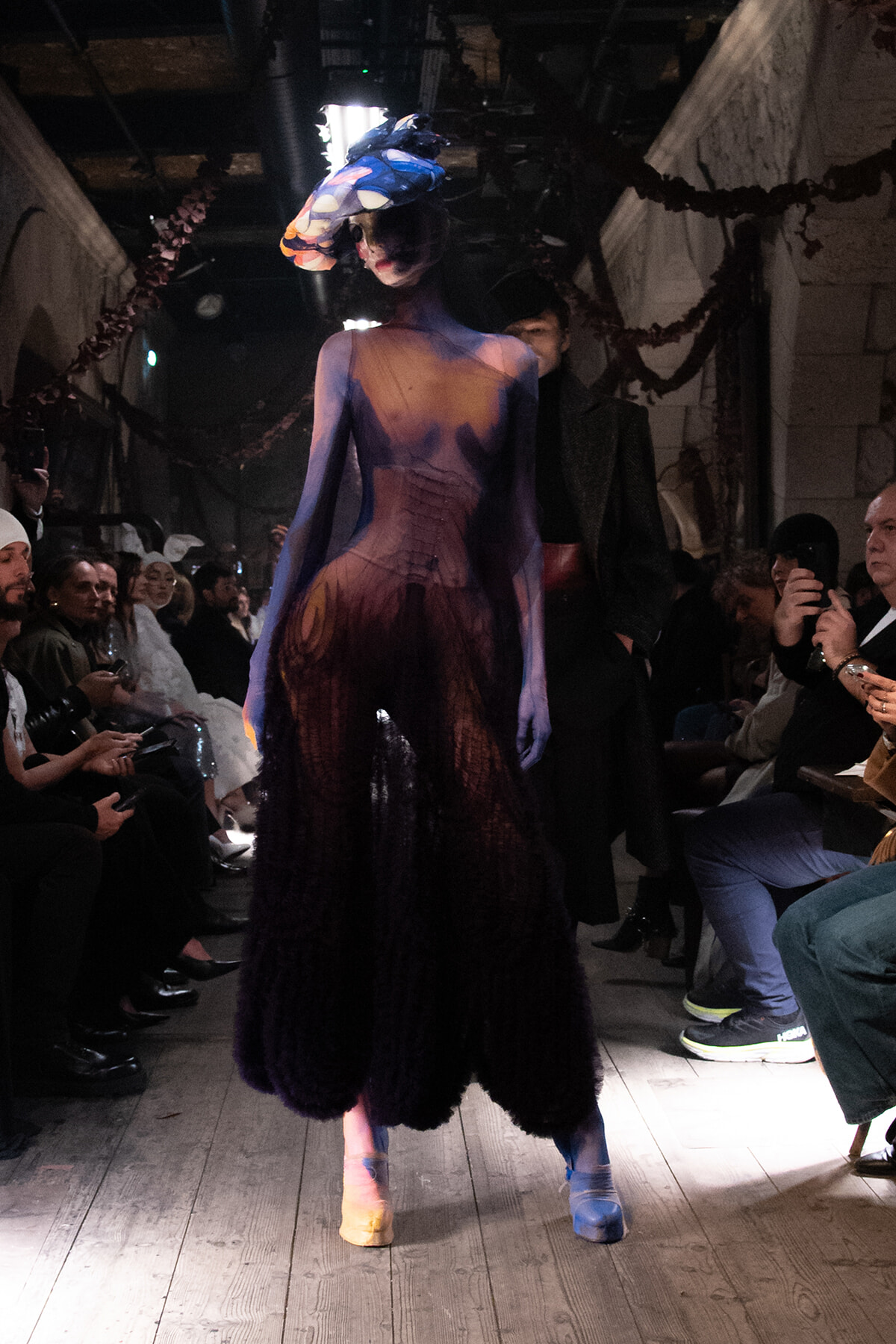 Model walks the runway in a gradient purple dress with a large colorful flower headpiece, audience watching from both sides.