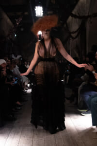 Model walks the runway in a sheer black gown with an oversized orange wig, audience seated along the catwalk in a dimly lit venue.
