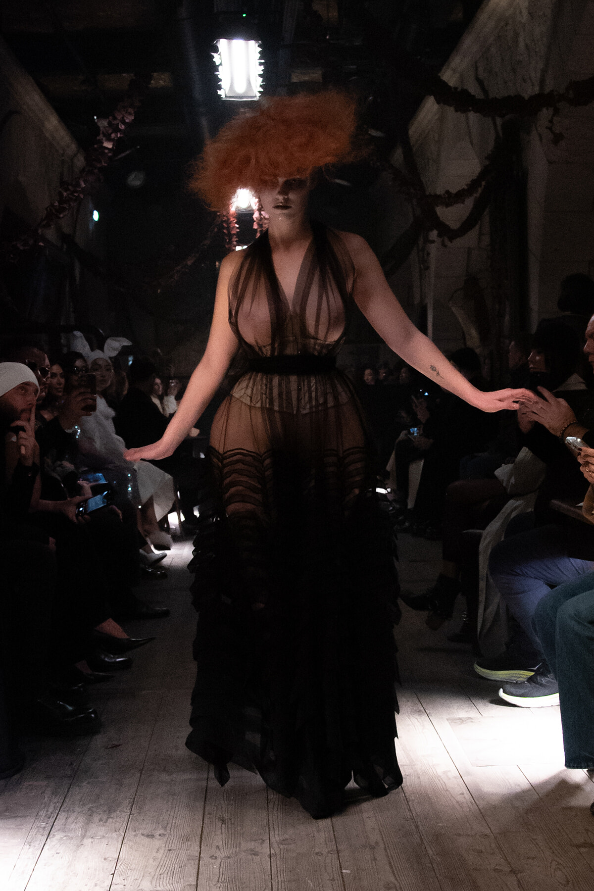 Model walks the runway in a sheer black gown with an oversized orange wig, audience seated along the catwalk in a dimly lit venue.