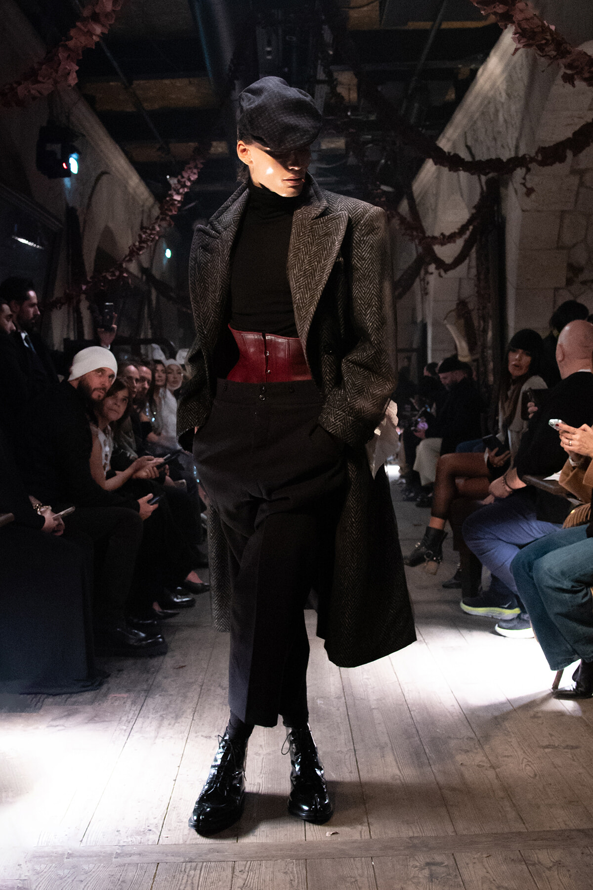 Model on a runway wearing a herringbone coat, black turtleneck, red waist belt, black pants, and shiny black boots; audience seated along the sides.