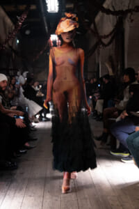 Model walking runway in a sheer, color-blocked gown with ruffled, ombré headdress; audience on both sides illuminated by spotlights.