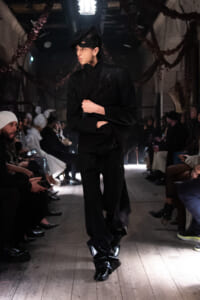 Model walks a dark runway in an all-black ensemble with a dramatic hat, audience seated on both sides.