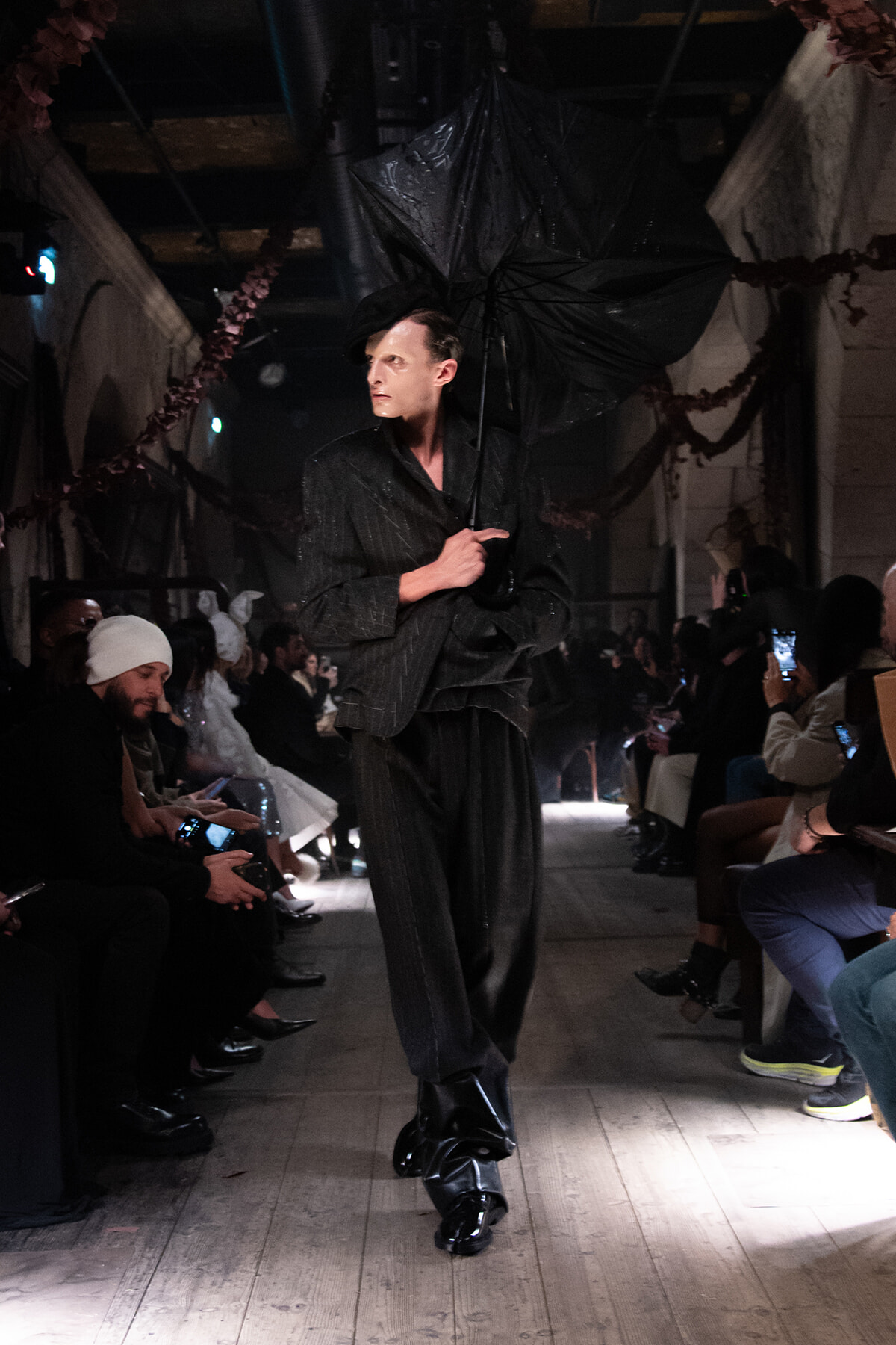 Model walking a runway in a dark pinstripe suit, holding a black umbrella, audience on both sides watching and photographing the show