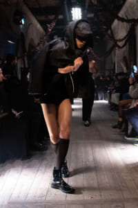 Runway model in a black hooded cape and cap, wearing shiny black boots, walking the dimly lit catwalk with audience on both sides.