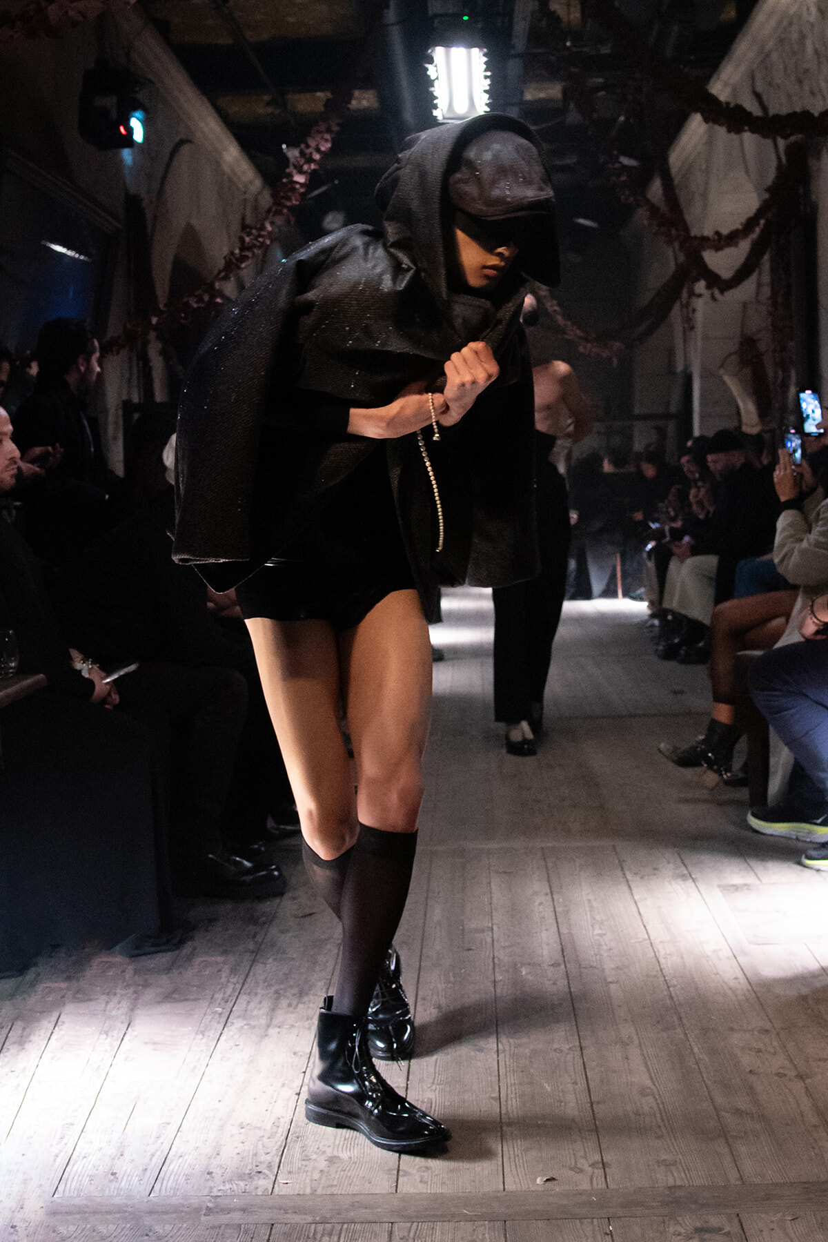 Runway model in a black hooded cape and cap, wearing shiny black boots, walking the dimly lit catwalk with audience on both sides.
