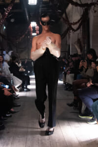 Male-presenting model walking a runway at a fashion show, shirtless with oversized white cuffed sleeves, black high-waisted pants, and dark sunglasses, as audience members photograph from the sides.