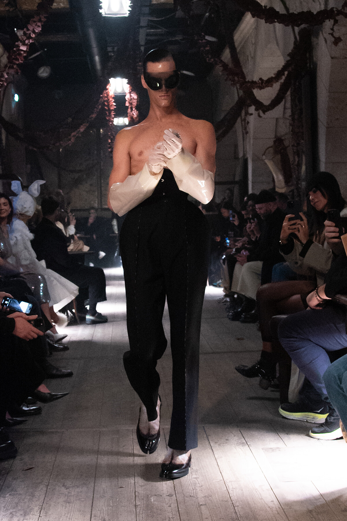 Male-presenting model walking a runway at a fashion show, shirtless with oversized white cuffed sleeves, black high-waisted pants, and dark sunglasses, as audience members photograph from the sides.