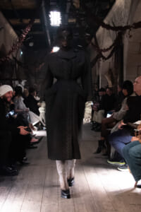 Runway fashion show with a tall model in a long black coat and white gloves, audience seated along both sides.