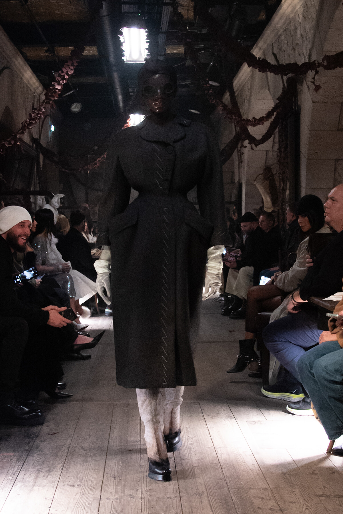 Runway fashion show with a tall model in a long black coat and white gloves, audience seated along both sides.