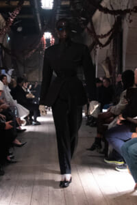 Model walks a dark, tailored black suit down a runway during a fashion show; audience seated along both sides in a dimly lit venue.