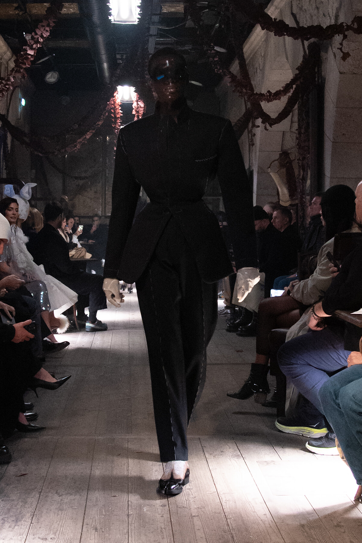 Model walks a dark, tailored black suit down a runway during a fashion show; audience seated along both sides in a dimly lit venue.