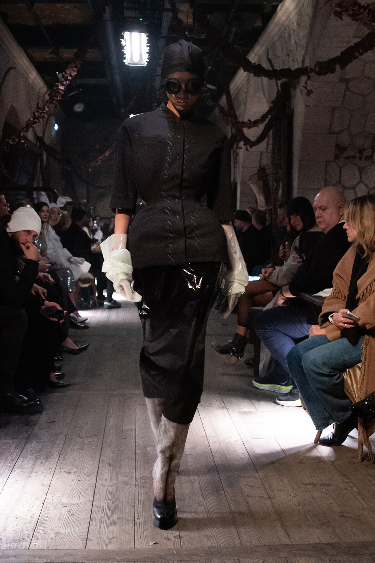 Fashion model walks a runway in a dark structured coat, oversized white gloves, and black patent-like pants in a dimly lit venue with seated audience nearby.