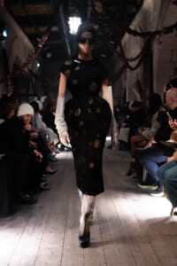 Model walks a runway in a black dress with circular cutouts, white satin gloves, round goggles, and a headwrap in a dim, spectator-filled venue.