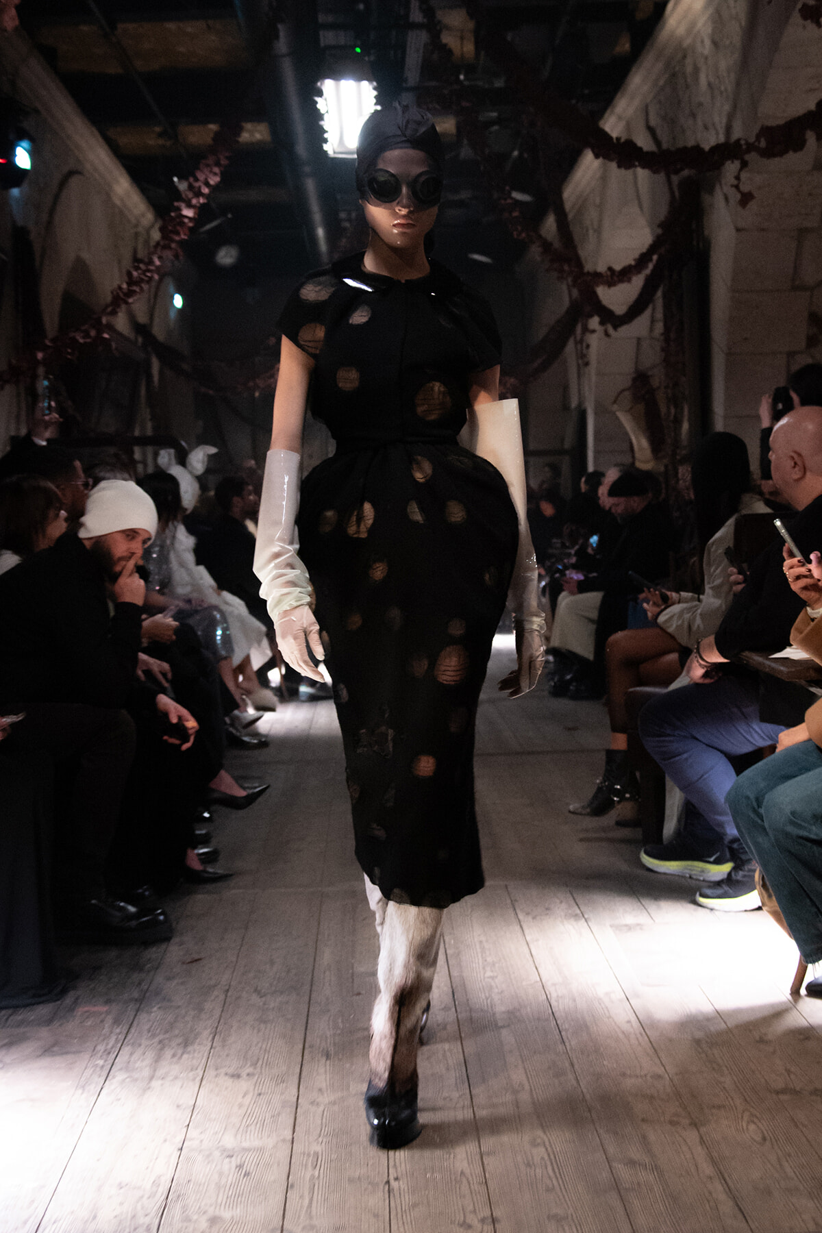 Model walks a runway in a black dress with circular cutouts, white satin gloves, round goggles, and a headwrap in a dim, spectator-filled venue.