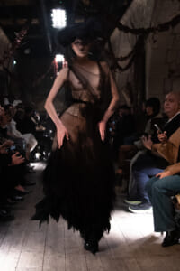 Model walks a dimly lit runway in a sheer black gown with a voluminous tulle skirt, audience seated along both sides watching.