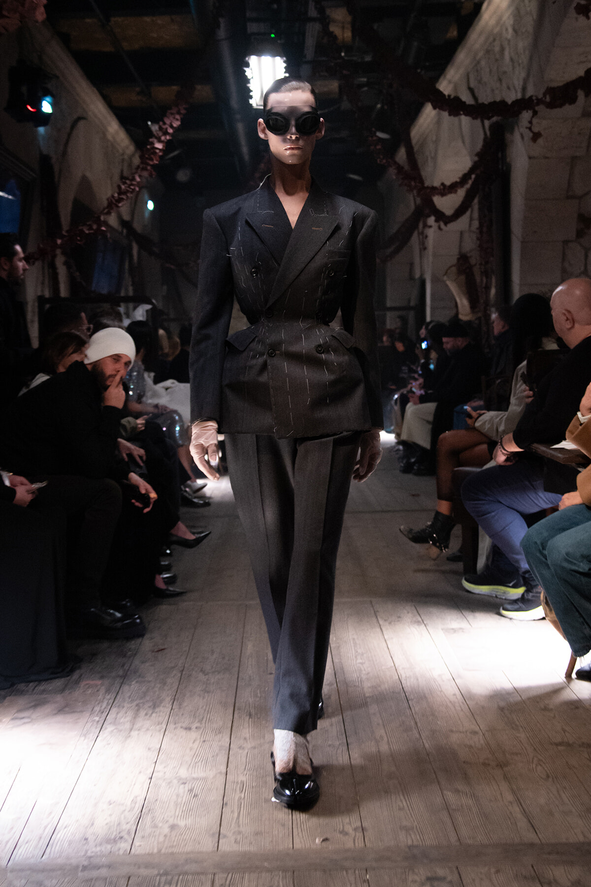 Model walking a wooden runway in a dark pinstripe suit with a double-breasted blazer and oversized goggles, audience on both sides.