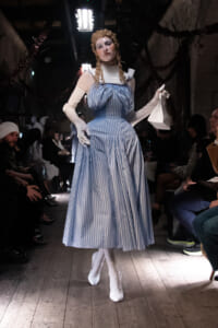 Model walking the runway in a blue and white striped ball gown with long white gloves, carrying a small white purse; audience seated on either side.