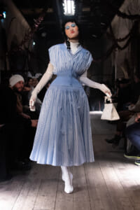 Model walking the runway in a blue striped midi dress with white gloves and white ankle boots, carrying a small white handbag.