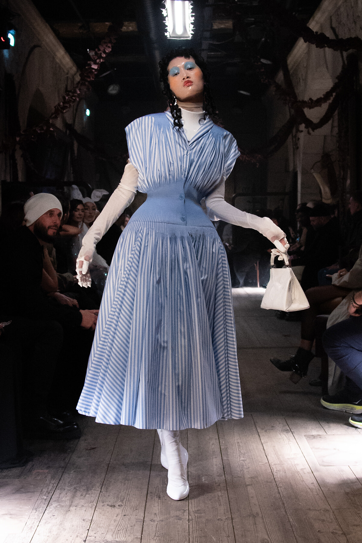 Model walking the runway in a blue striped midi dress with white gloves and white ankle boots, carrying a small white handbag.
